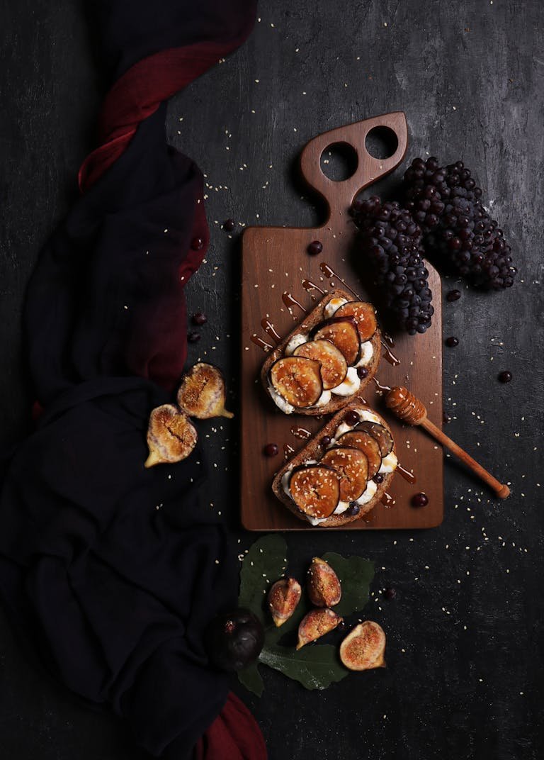 Dark, artistic flat lay of fig and grape toast with honey on a rustic cutting board.