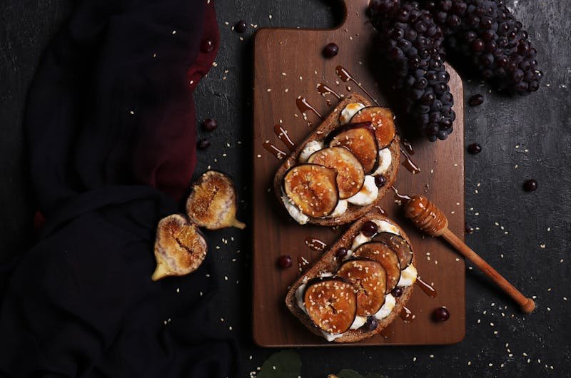 Dark, artistic flat lay of fig and grape toast with honey on a rustic cutting board.