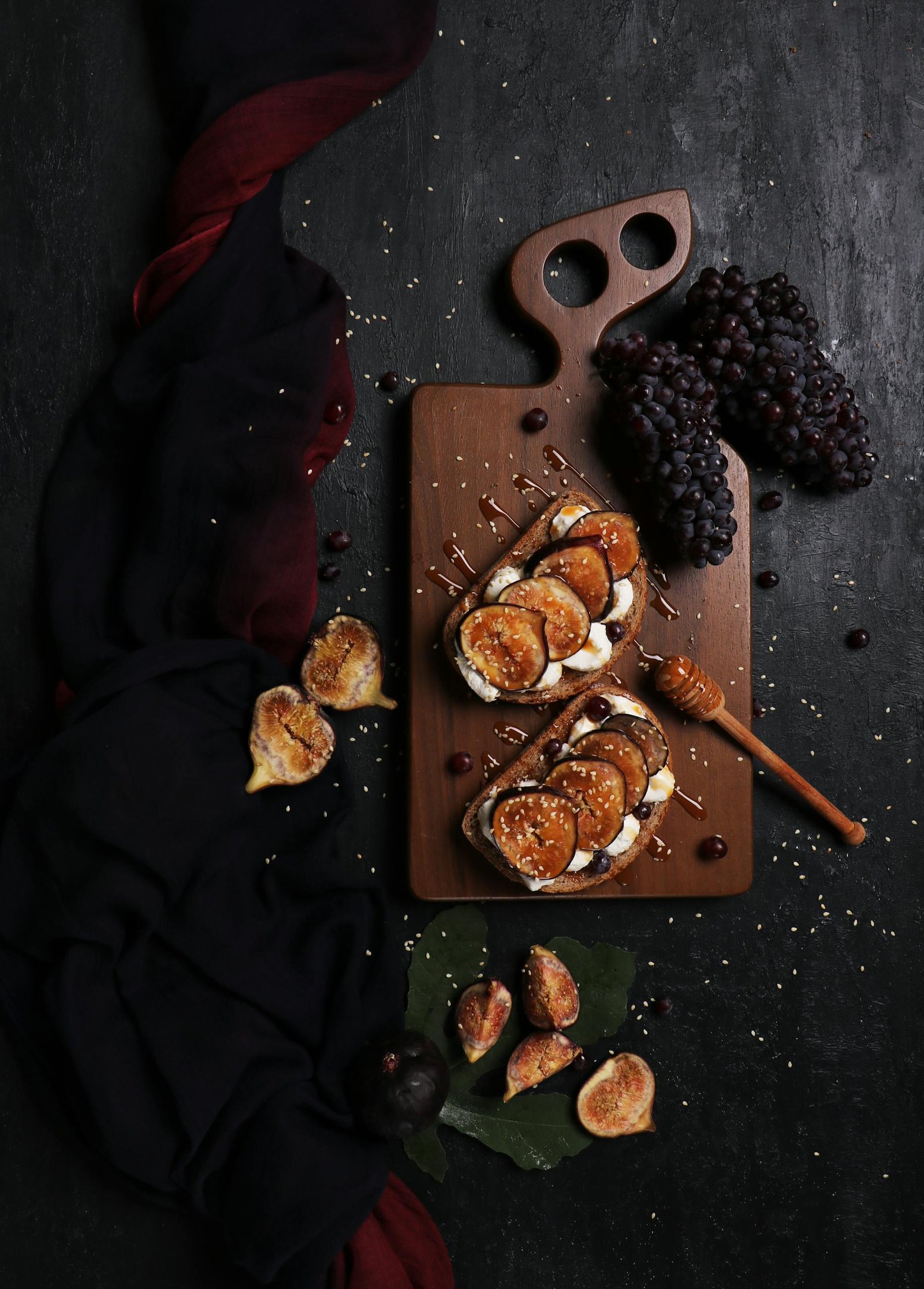 Dark, artistic flat lay of fig and grape toast with honey on a rustic cutting board.